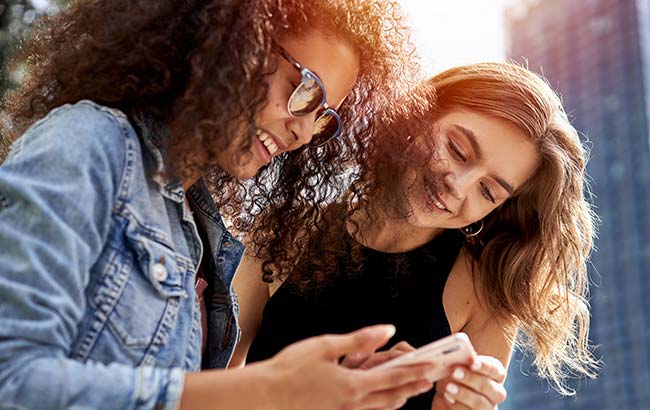 Two young women smiling while looking at social media content on a smartphone outdoors